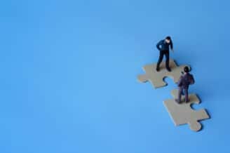 Two miniature figures representing businessmen stand on puzzle pieces symbolizing Leadfeeder merger and rebrand to Dealfront