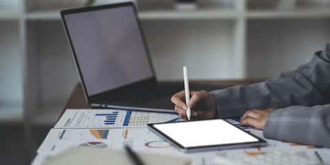 businesswoman's hand holding a pen with papers and a tablet in front of her on a desk representing sales intelligence tools