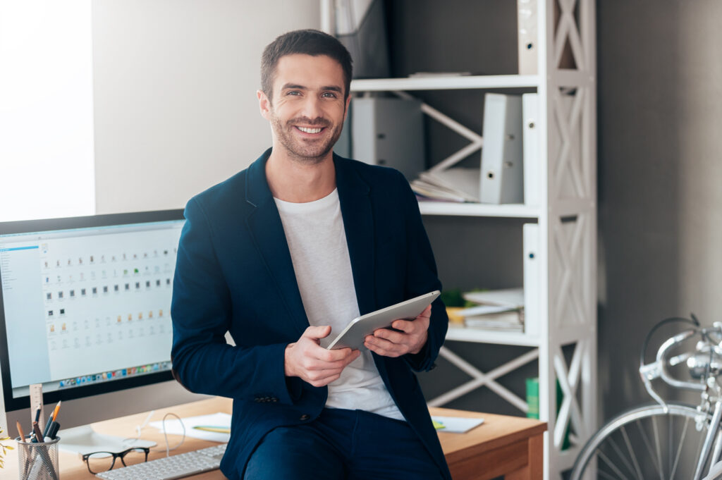 Businessman stands in front of his desk holding a tablet and looking at the camera as he thinks about sales techniques