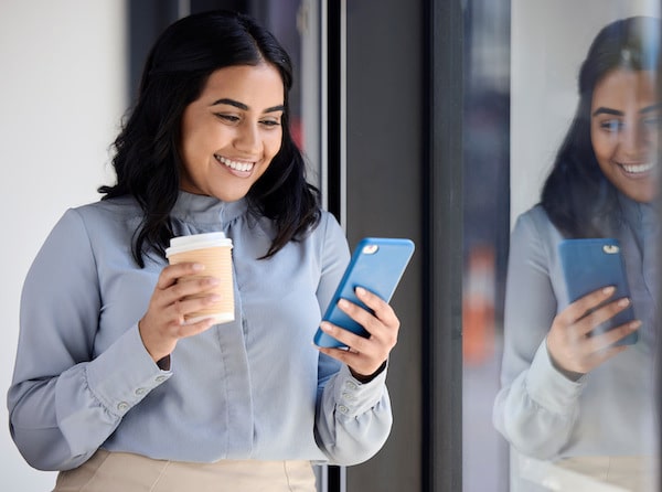 Young business woman stands holding a coffee cup and her cellphone smiling as she participates in social selling