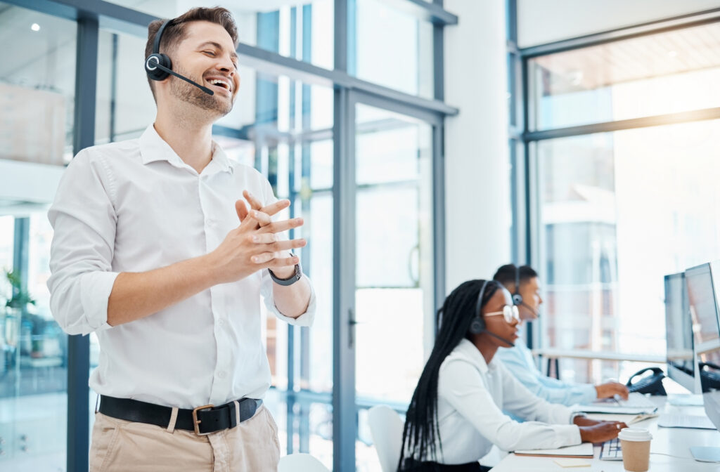 Sales rep with headset on stands in office talking on phone as part of his social selling strategy, with two other reps in the background