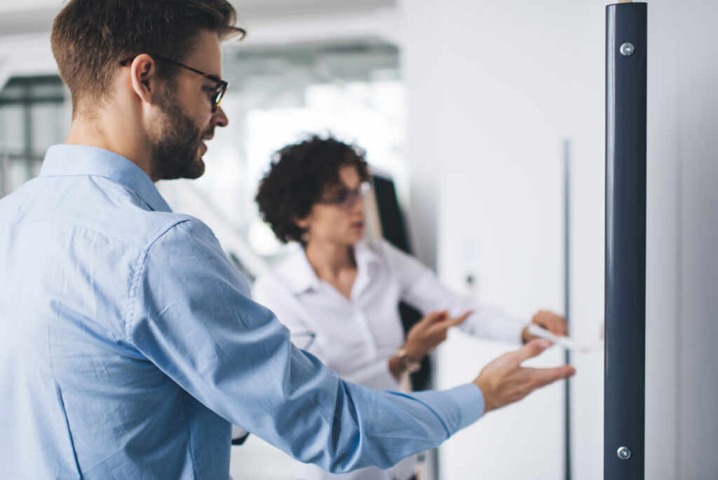 Two business professionals stand at interactive display board brainstorming together, symbolizing the benefits of in-person work