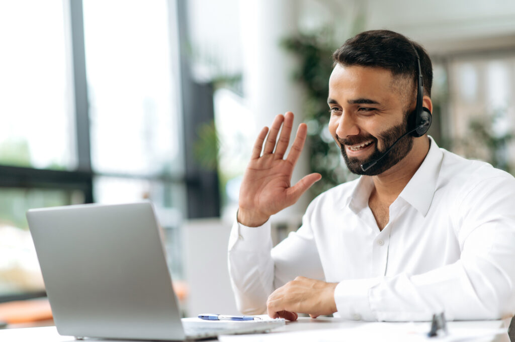 Sales rep on sitting in front of laptop with headset on waving to prospect on a video call representing outbound sales