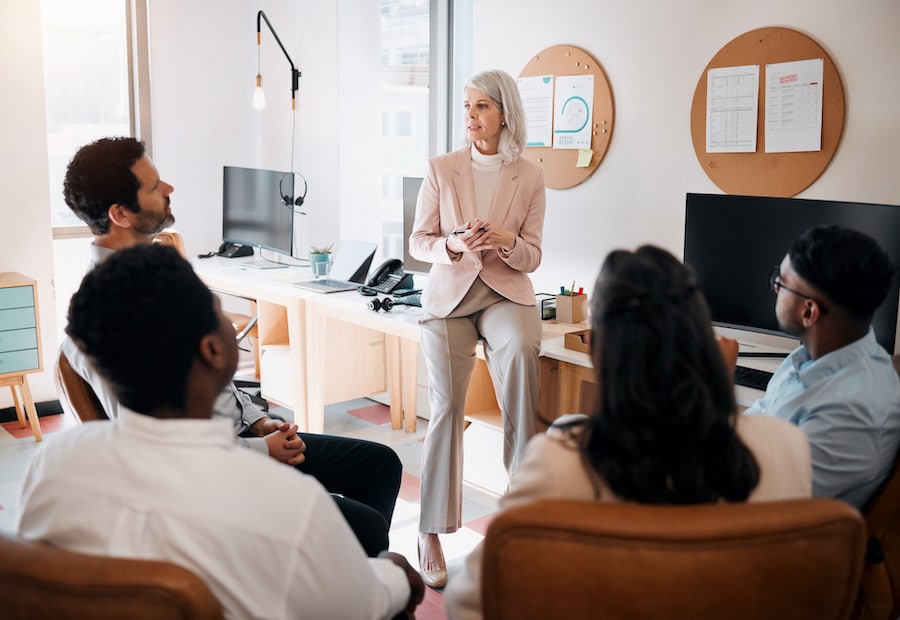 group of business professionals sit in a circle looking at an older business woman presenting a sales data training to them