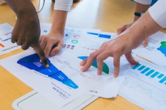 The hands of three business people point at graphs and charts on a table, symbolizing the B2B sales funnel