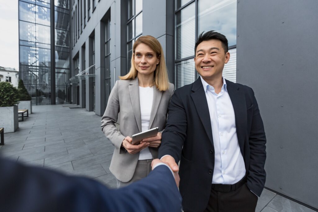 A business man and woman stand facing another man, the two business men shake hands representing B2B sales during a recession