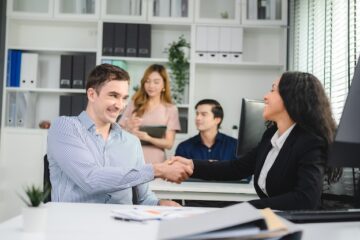 Two sales professionals sit at desks next to each other, shaking hands in celebration of a sales win