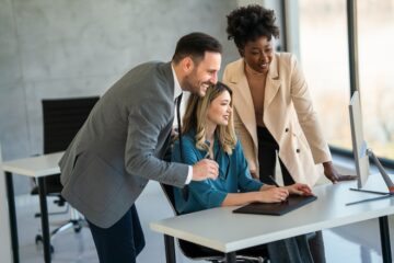 A group of sales professionals stands around a co-worker sitting at a desk looking at a computer reviewing their B2B sales automation plan