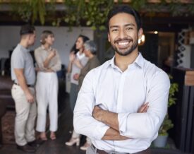 A business man stands facing the camera smiling with colleagues standing in the background