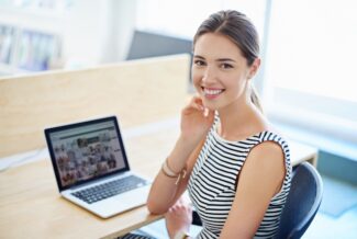 Female business professional sits at a desk with her laptop in front of her, looking at camera and smiling as she thinks about B2B personalization