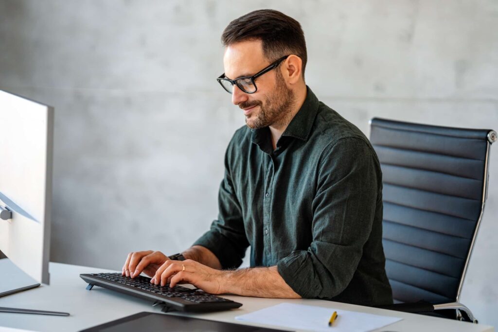 Business man sits at desk in front of a desktop computer, typing tailored outreach as part of a B2B personalization strategy